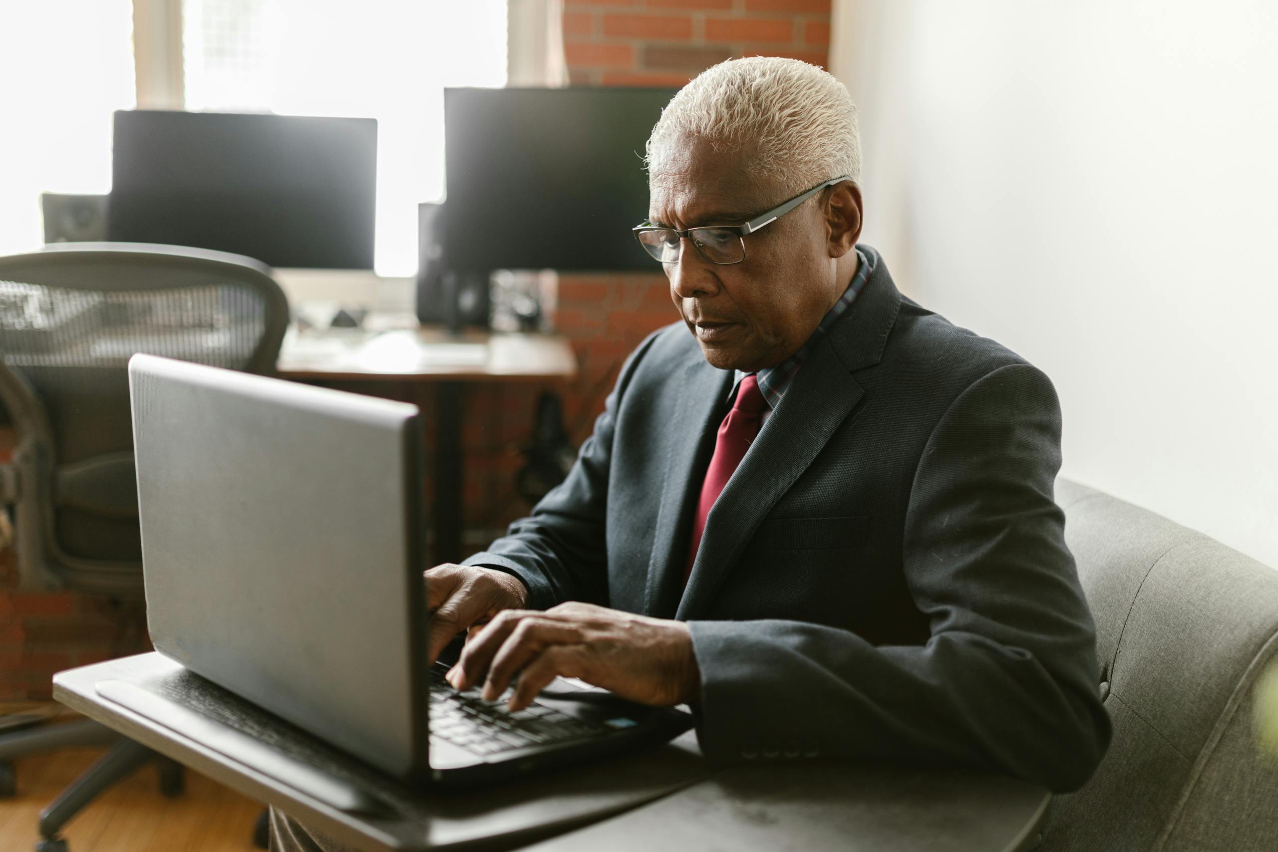 Elderly man in business attire using a laptop in a modern office setting.