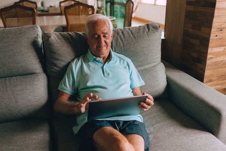 Older man using tablet on a sofa in cozy living room, enjoying leisure time indoors.